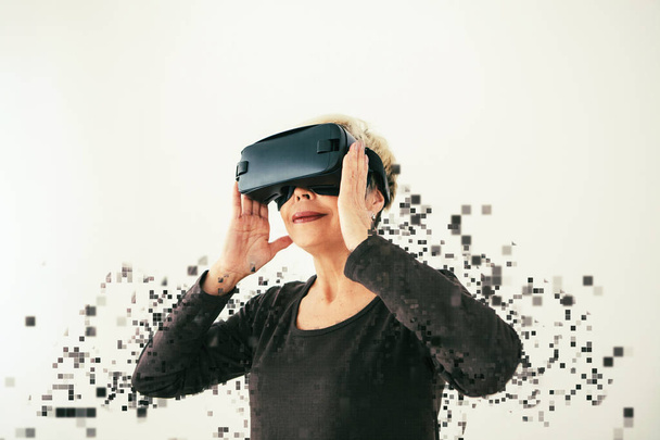 An elderly woman in virtual reality glasses is scattered by pixels. Conceptual photography with visual effects with an elderly person using modern technology. - Photo, Image