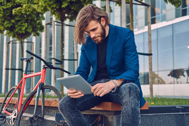 Μοντέρνα όμορφος γενειοφόρος άνδρας με μακριά μαλλιά που κάθεται σε ένα παγκάκι με ένα ποδήλατο, χρησιμοποιώντας έναν υπολογιστή tablet. - Φωτογραφία, εικόνα