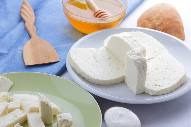 Μοτίβο τυρί, μέλι και ξινή κρέμα, φέτα σε λευκό και πράσινο πλάκα, μαλακό τυρί σε λευκό φόντο, ξύλινο κουτάλι με την ξινή κρέμα, μπλε πετσέτα, μέλι στα γυαλικά, τέχνη - Φωτογραφία, εικόνα