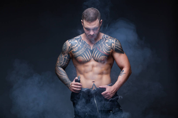 Vaper. The man with a muscular naked torso with tattoos smoke an electronic cigarette on the dark background - Foto, Imagem