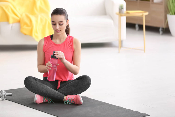 Mooie jonge vrouw met flesje water na het doen van fitness oefeningen thuis - Foto, afbeelding