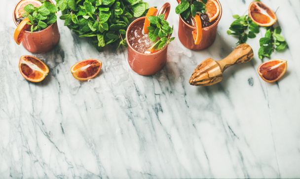 Flat-lay of blood orange Moscow mule alcohol cocktails with fresh mint and ice in copper mugs over white marble background, top view, copy space - Фото, изображение