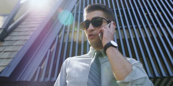 handsome businessman in sunglasses holding and using smart phone outdoor on modern building background - Imágenes, Vídeo