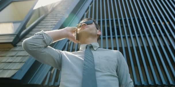 handsome businessman in sunglasses holding and using smart phone outdoor on modern building background - Felvétel, videó
