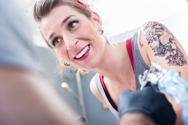 Retrato de una mujer alegre y de moda sonriendo mientras mira al artista del tatuaje con confianza en un estudio moderno con equipo profesional estéril
 - Foto, Imagen