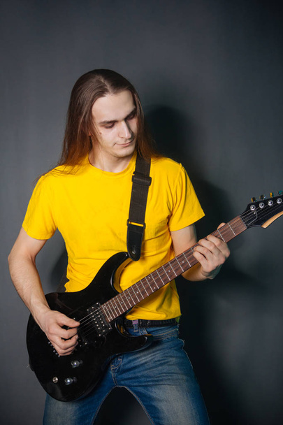 Hombre con el pelo largo en camiseta amarilla, tocando en la guitarra eléctrica rock. Retrato de estudio de moda sobre fondo gris
. - Foto, Imagen