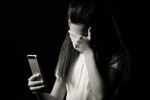 Single sad teen holding a mobile phone lamenting sitting on the bed in her bedroom with a dark light in the background - Photo, Image