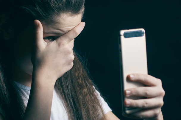 Μία θλιβερή έφηβος κρατώντας ένα κινητό τηλέφωνο, θρηνώντας κάθεται στο κρεβάτι στην κρεβατοκάμαρά της με ένα σκούρο φως στο παρασκήνιο - Φωτογραφία, εικόνα