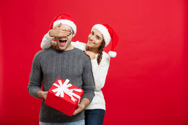Christmas Concept - Young woman covering mans eyes with hand and giving surprise big gift. Isolated on Red background - Photo, Image