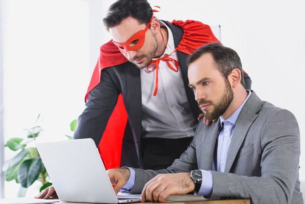 супер бизнесмен в маске и плаще помогает бизнесмену с работой в офисе
  - Фото, изображение