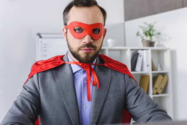 Porträt eines Superunternehmers in Maske und Umhang, der im Büro in die Kamera blickt  - Foto, Bild