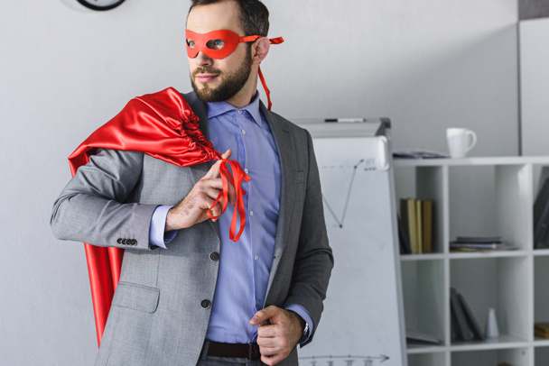 super businessman in mask holding cape on shoulder and looking away in office  - Zdjęcie, obraz