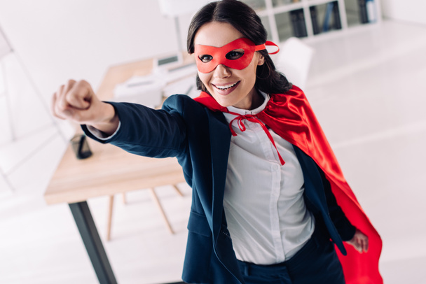 aantrekkelijke super zakenvrouw in cape en masker permanent met hand omhoog in office - Foto, afbeelding