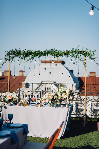 Closeup of festive composition with flowers in golden vases, white candles and tableware on background of wedding arch with branches and lamps - Photo, Image