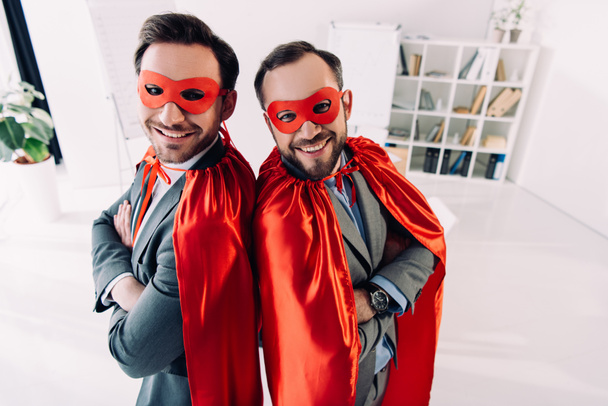 visão de alto ângulo de sorrir bonitos super empresários em máscaras e capas com braços cruzados no escritório
 - Foto, Imagem