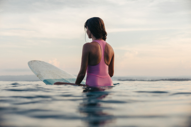 back view of girl sitting on surfboard in ocean at sunset - Фото, изображение