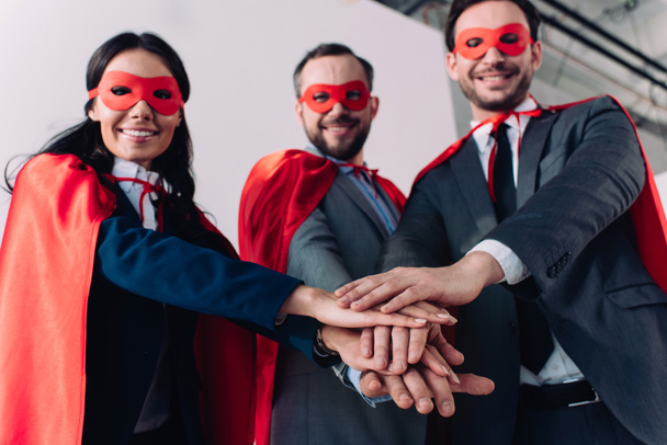 visão de baixo ângulo de super empresários em máscaras e capas colocando as mãos juntas no escritório
 - Foto, Imagem