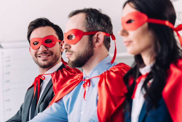 Seitenansicht lächelnder Superunternehmer in Masken und Umhängen, die im Büro wegschauen - Foto, Bild