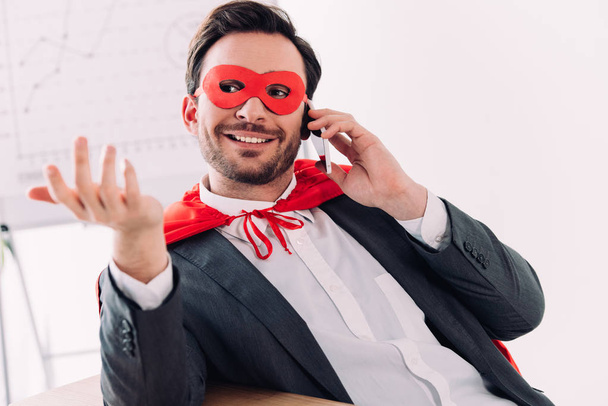 handsome super businessman in mask and cape talking by smartphone in office - Photo, Image