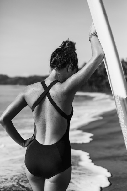 back view of young girl in swimsuit posing with surfboard near ocean, black and white - Photo, Image