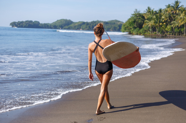 Achteraanzicht van jonge vrouwelijke surfer wandelen met surfboard op strand - Foto, afbeelding
