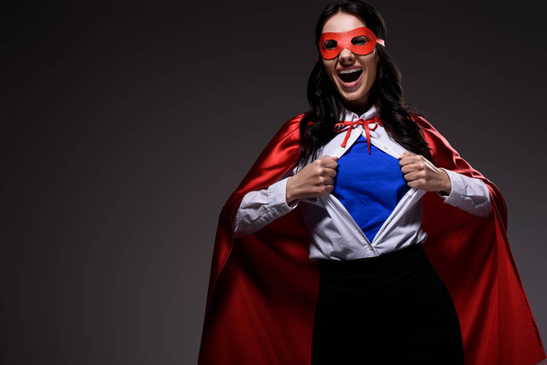 lachen aantrekkelijke super zakenvrouw in rode cape en masker weergegeven: blauw shirt geïsoleerd op zwart - Foto, afbeelding