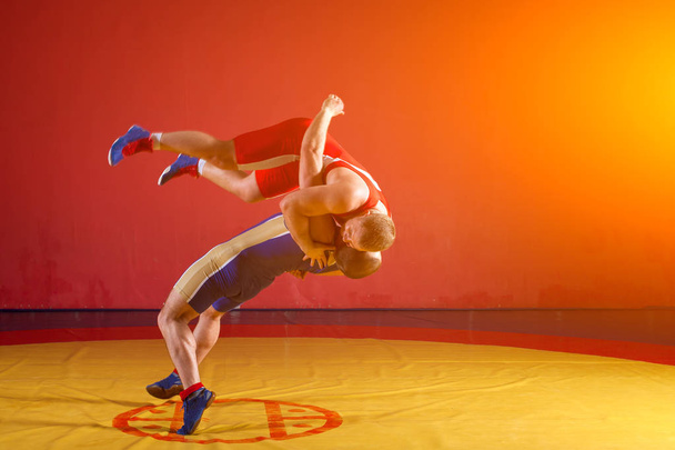 Two young men in blue and red wrestling tights are wrestlng and making a suplex wrestling on a yellow wrestling carpet in the gym - Photo, Image