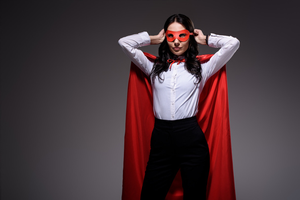 attractive super businesswoman in red cape tying mask isolated on black - Fotografie, Obrázek