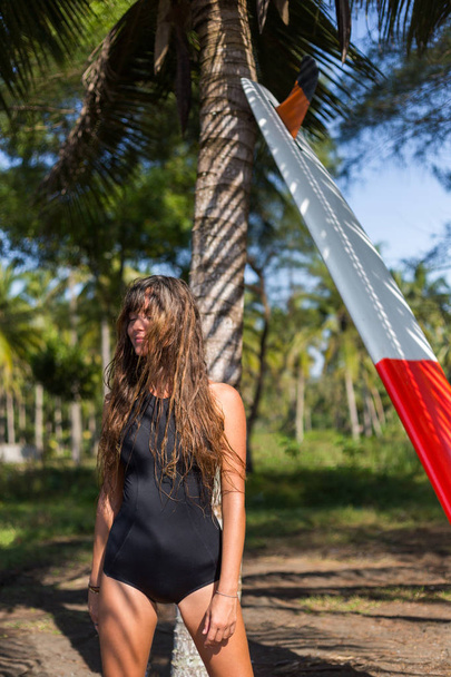 young woman with long hair posing with surfboard near palm tree - Foto, imagen