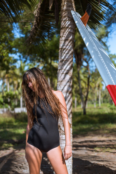 female surfer in black swimsuit posing with surfboard near palm tree - Foto, immagini