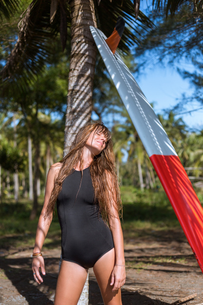 smiling tanned girl posing in black swimsuit with surfboard near palm - Fotografie, Obrázek