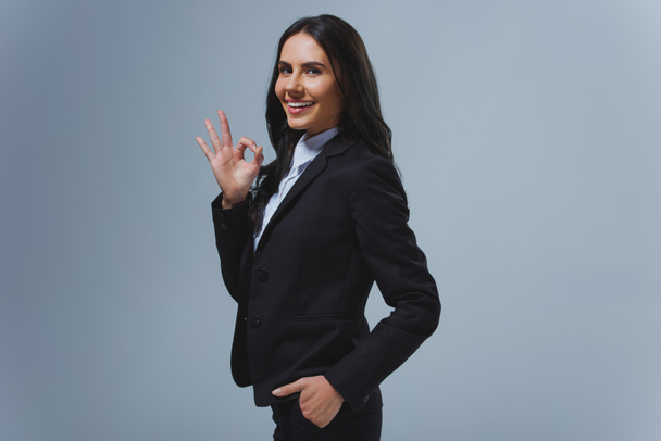 smiling attractive businesswoman showing okay gesture isolated on grey - Photo, Image