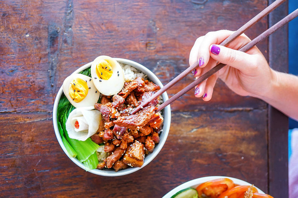 Μπολ ρυζιού σιγοβρασμένο κοιλιά χοιρινό κρέας σε ένα ξύλινο τραπέζι με αυγό και σαλάτα. Ένα χέρι με chopsticks κατέχει ένα κομμάτι κρέας. Το Top view με αντίγραφο χώρου - Φωτογραφία, εικόνα