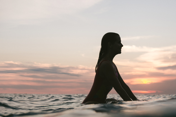 vista lateral de la silueta de la mujer descansando sobre tabla de surf en el océano al atardecer
 - Foto, imagen