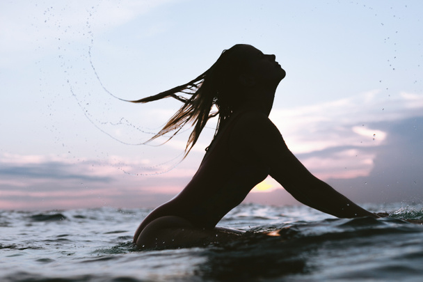silhouette of woman whipping hair while resting on surfing board in ocean on sunset - Photo, Image