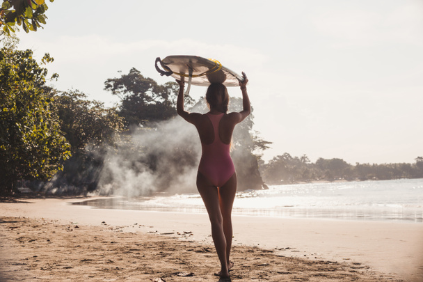 Achteraanzicht van Sportvrouw surfen Bestuur dragen op hoofd tijdens het wandelen op de kustlijn - Foto, afbeelding