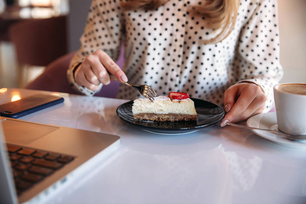 Girl eating cake in a cafe. Blonde in polka-dot shirt with laptop - Fotoğraf, Görsel