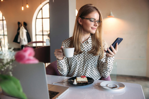 Una chica comiendo pastel en un café. Rubia en camisa de lunares con portátil
 - Foto, imagen