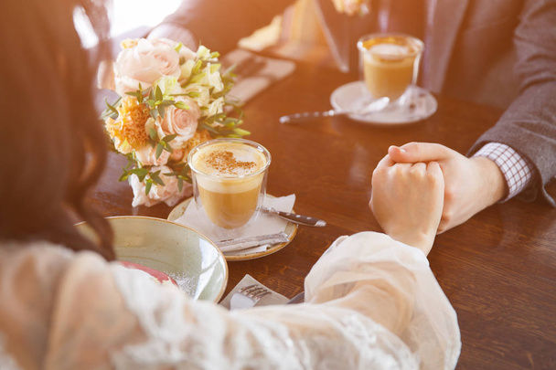 Νύφη και γαμπρός σε ένα πολυτελές εστιατόριο, κρατώντας τα χέρια και πίνοντας ένα φλιτζάνι καφέ latte. - Φωτογραφία, εικόνα