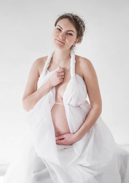 φωτογραφία έγκυος κορίτσι με ένα διάδημα σε μια λευκή κουρτίνα σε λευκό φόντο με το χέρι στην καρδιά - Φωτογραφία, εικόνα