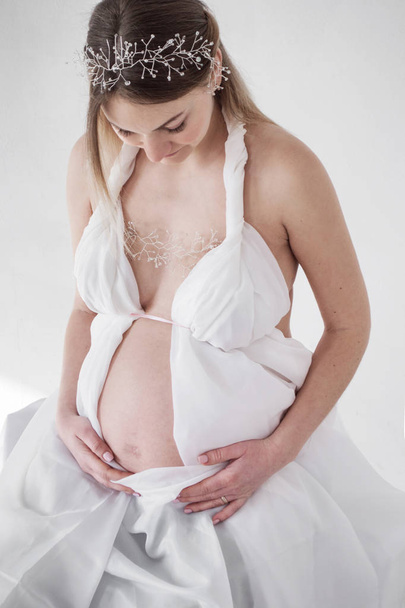 Dziewczyna w ciąży zdjęcia w białe szaty na białym tle ręce na brzuchu - Zdjęcie, obraz