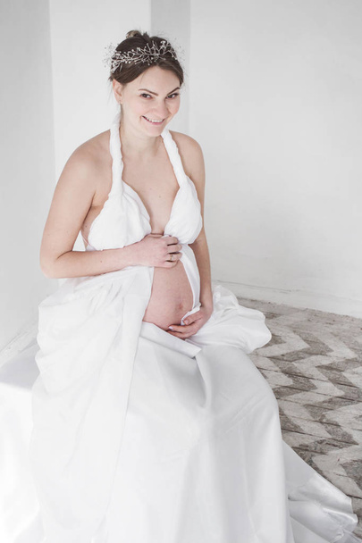 foto Menina grávida sorrindo em um diadema em um cortinado branco em um fundo branco senta-se
 - Foto, Imagem