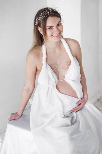 foto Mulher grávida sorrindo em uma tiara em cortinas brancas senta-se em um fundo branco
 - Foto, Imagem