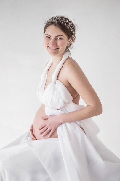 白い背景に白いドレスで笑って写真妊婦が彼女の胃に手します。 - 写真・画像