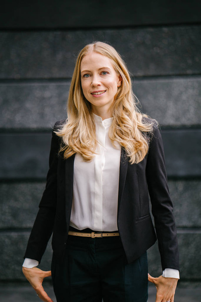 Portrait of a Caucasian, white and attractive middle-aged woman in a professional suit against a dark grey background. She is beautiful with blond hair and blue eyes and smiling  - Foto, Bild