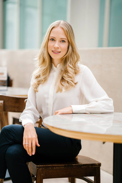 Portrait of a young, Caucasian European woman sitting at a table during the day in an office with lots of natural light. She is blond, has blue eyes, and is professionally dressed and is smiling - Foto, Imagem
