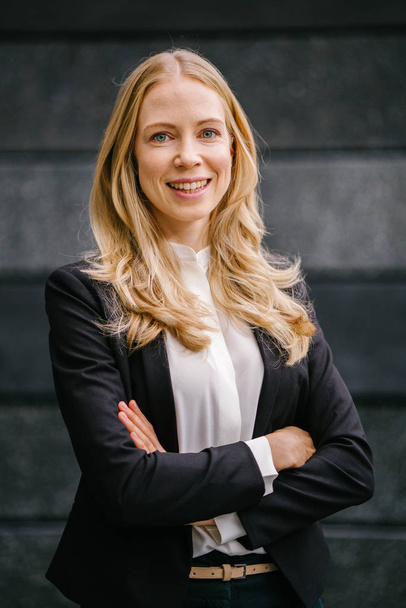 Portrait of a Caucasian, white and attractive middle-aged woman in a professional suit against a dark grey background. She is beautiful with blond hair and blue eyes and smiling  - Фото, зображення