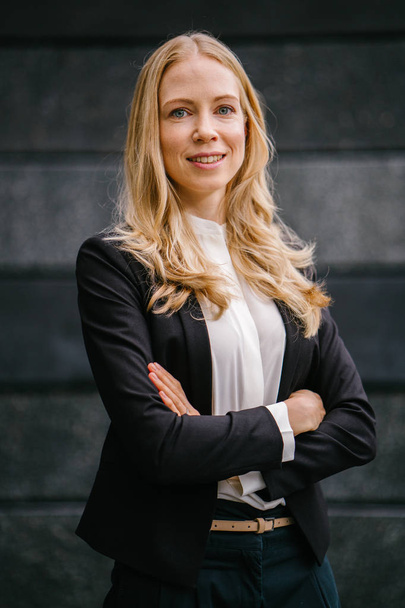 Portrait of a Caucasian, white and attractive middle-aged woman in a professional suit against a dark grey background. She is beautiful with blond hair and blue eyes and smiling  - Fotó, kép