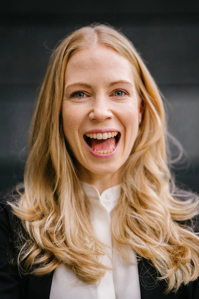 Portrait of a Caucasian, white and attractive middle-aged woman in a professional suit against a dark grey background. She is beautiful with blond hair and blue eyes and smiling for her head shot. - 写真・画像