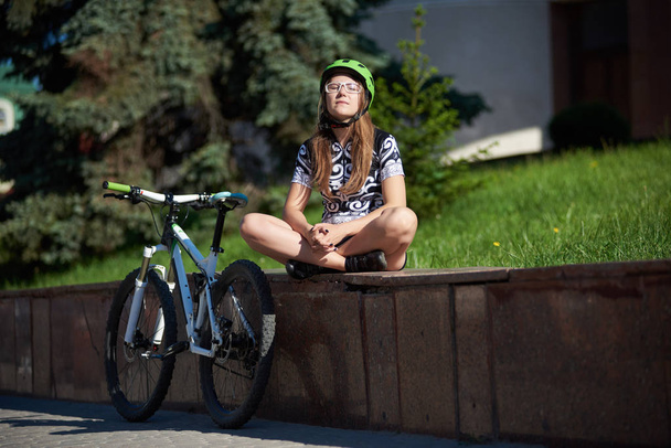 目で太陽を楽しむプロフェッショナル サイクリング ウエアとヘルメット自転車、近く通り縁の上に座って女性サイクリストの閉鎖. - 写真・画像